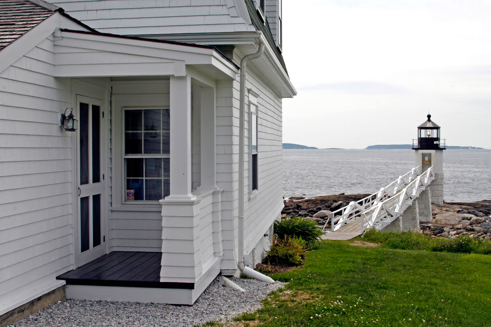 The porch of a white timber house with a walkway leading to a lighthouse. 