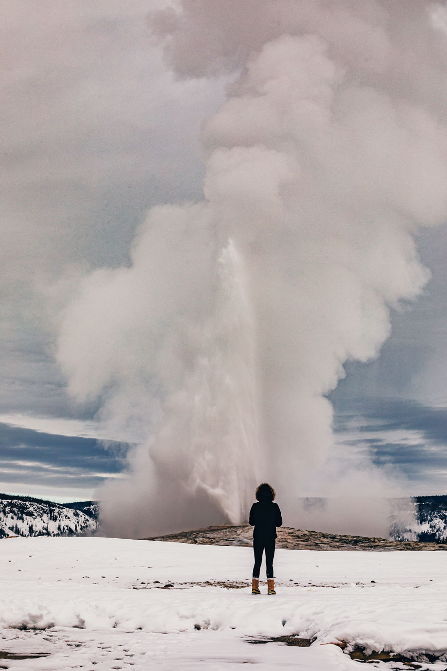 A woman stands in front of an erupting geyser.