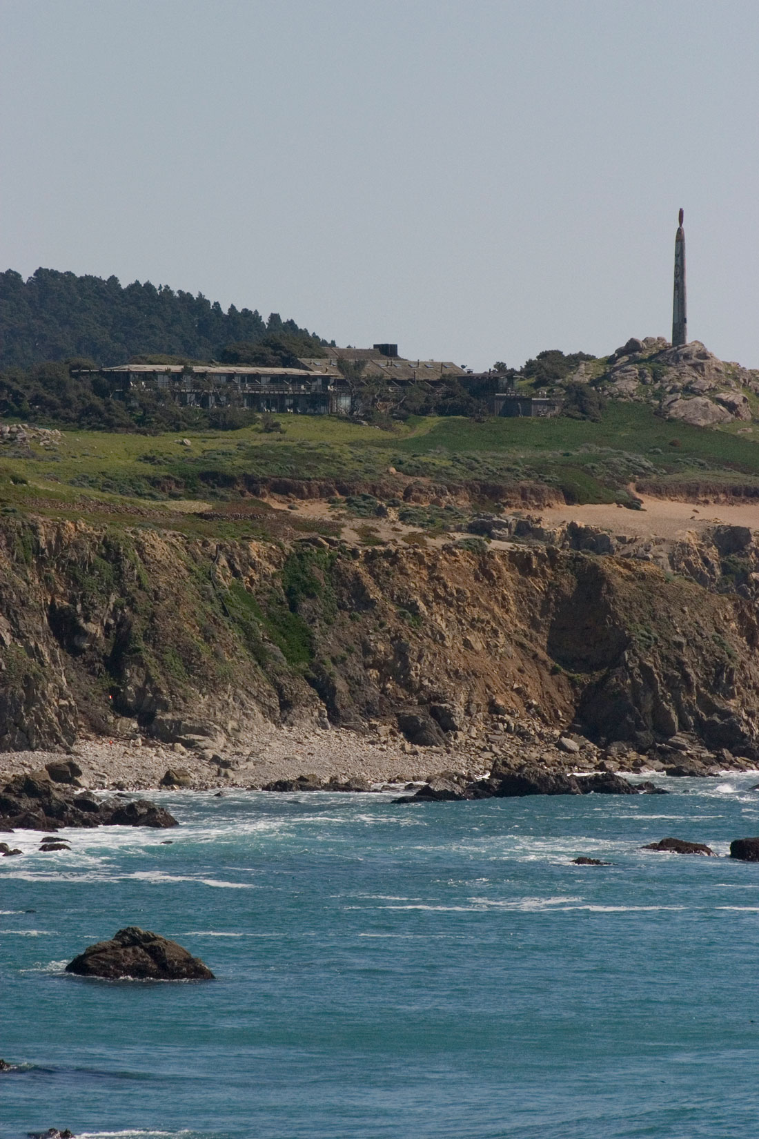 A wooden hotel and an obelisk sit on ground above cliffs leading down to the ocean. 