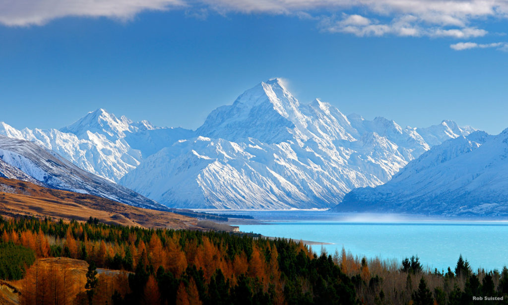 A snowy mountain landscape next to an ice blue lake in autumn colours.