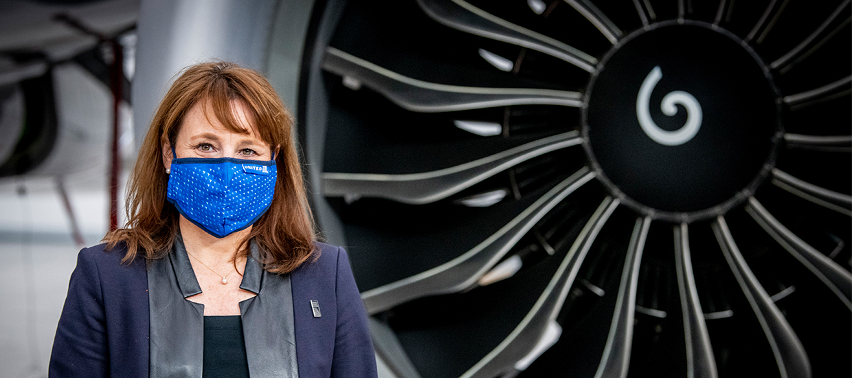 Red haired woman stands in front of airplane jet wearing navy blazer with black lapels and a bright blue face mask